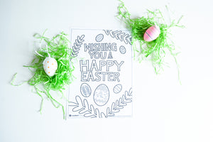 A coloring sheet on a white tabletop. There’s fake Easter grass around the color page. The page features the words “Wishing you a happy Easter” with illustrated Easter eggs and palm leaves.