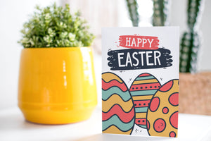 A greeting card is on a table top with a yellow plant pot and a green plant inside. The card features the words “Happy Easter” with illustrated Easter eggs in muted bright colors. 