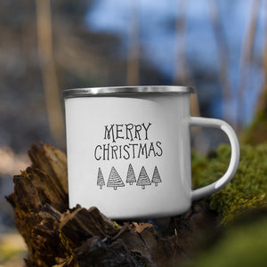 White enamel mug sitting on top of a tree branch with moss in the background. The design on the mug is featured in black reading "Merry Christmas" with modern illustrated trees below the words. 