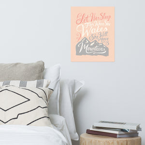 A pink print hanging on a pale grey wall, over a white bed with grey and white cushions. The print reads 'Let her sleep for when she wakes she will move mountains' in a pink, white, and light grey lettering design, with a grey mountain illustration at the bottom.