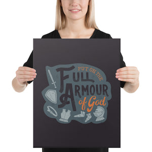 A smiling woman holds up a dark gray canvas. The canvas features the quote 'Put on the full armour of God' in black and orange typography, with medieval-style pieces of armour illustrated in pale gray.
