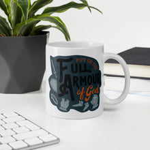 Load image into Gallery viewer, A white mug sits on a white desk beside a keyboard, notebook, and potted plant. The mug features the quote &#39;Put on the full armour of God&#39; in black and orange typography, along with illustrated pieces of armour in medieval style against a dark gray background.