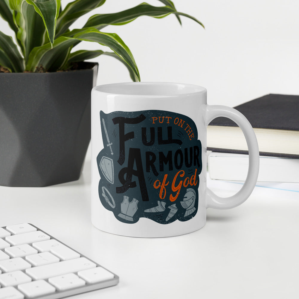 A white mug sits on a white desk beside a keyboard, notebook, and potted plant. The mug features the quote 'Put on the full armour of God' in black and orange typography, along with illustrated pieces of armour in medieval style against a dark gray background.