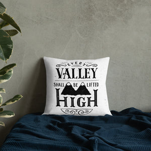 A monochrome square pillow sits on a blue bedspread against a grey concrete wall. The pillow design is black on a white background, and reads 'Every valley shall be lifted high' in a variety of typographic lettering, with flourishes and an illustration of two mountain peaks. 