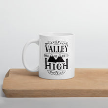 Load image into Gallery viewer, A white mug sits on a wooden board against a white backdrop. The mug has a typographic design in black text, with the words &#39;Every valley shall be lifted high&#39; lettered with flourishes and an illustration of two mountain peaks.