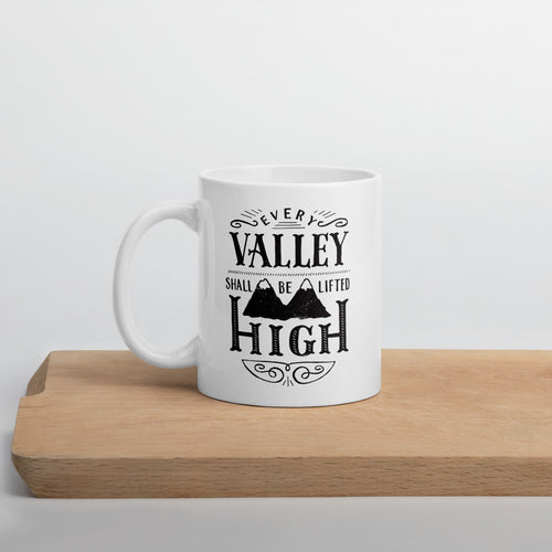 A white mug sits on a wooden board against a white backdrop. The mug has a typographic design in black text, with the words 'Every valley shall be lifted high' lettered with flourishes and an illustration of two mountain peaks.