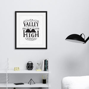 A monochrome black and white print in a black frame hangs on a living room wall. The print reads 'Every valley shall be lifted high' in a variety of typographic lettering, with flourishes and an image of two mountain peaks. The living room is also monochrome with pale walls and a black lamp and accessories.