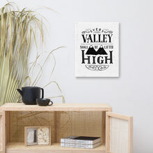 Load image into Gallery viewer, A monochrome art canvas hangs on a white wall. The canvas has a white background, with the words &#39;Every valley shall be lifted high&#39; in black typographic lettering with flourishes and an image of two mountain peaks. Beneath the canvas there’s a rattan cabinet holding black crockery and magazines.