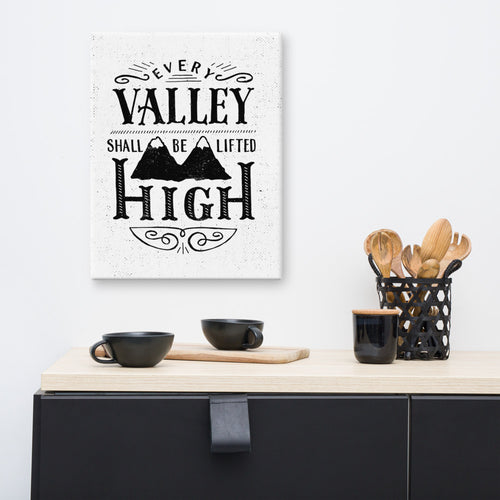 A monochrome art canvas hangs on a white kitchen wall. The canvas has a white background, with the words 'Every valley shall be lifted high' in black typographic lettering with flourishes and an image of two mountain peaks. Beneath the canvas there’s a black kitchen cabinet with a mixture of black crockery and wooden kitchen implements on top.