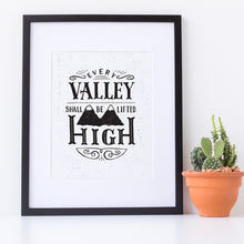 Load image into Gallery viewer, A small monochrome black and white print in a black frame sits against a white wall. The print reads &#39;Every valley shall be lifted high&#39; in a variety of typographic lettering, with flourishes and an image of two mountain peaks. Next to the print is a small potted cactus.