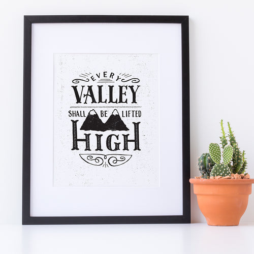 A small monochrome black and white print in a black frame sits against a white wall. The print reads 'Every valley shall be lifted high' in a variety of typographic lettering, with flourishes and an image of two mountain peaks. Next to the print is a small potted cactus.