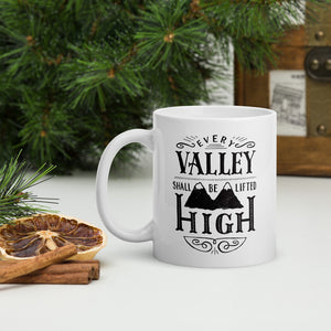 A white mug sits on a white worktop surrounded by evergreen foliage and cinnamon sticks. The mug has a typographic design in black text, with the words 'Every valley shall be lifted high' lettered with flourishes and an illustration of two mountain peaks.