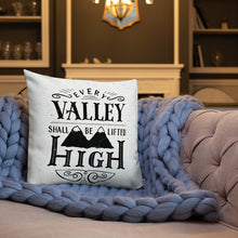 Load image into Gallery viewer, A monochrome square pillow sits on a comfortable sofa covered with a chunky-knit purple throw. The pillow design is black on a white background, and reads &#39;Every valley shall be lifted high&#39; in a variety of typographic lettering, with flourishes and an illustration of two mountain peaks. In the background is a dark coloured living room with bookshelves and wall lights.