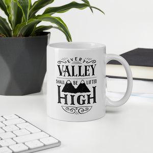 A white mug sits on a white desk surrounded by a computer keyboard, notebooks, and a potted plant. The mug has a typographic design in black text, with the words 'Every valley shall be lifted high' lettered with flourishes and an illustration of two mountain peaks.