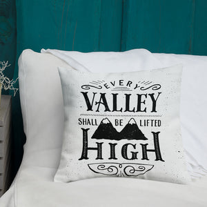 A monochrome square pillow sits on a white sofa against a teal fence. The pillow design is black on a white background, and reads 'Every valley shall be lifted high' in a variety of typographic lettering, with flourishes and an illustration of two mountain peaks. 