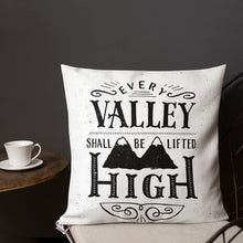 Load image into Gallery viewer, A monochrome square pillow sits on a chair against a dark grey wall. The pillow design is black on a white background, and reads &#39;Every valley shall be lifted high&#39; in a variety of typographic lettering, with flourishes and an illustration of two mountain peaks. Next to the chair is a small table with a white cup and saucer on it.