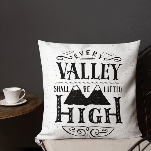 A monochrome square pillow sits on a chair against a dark grey wall. The pillow design is black on a white background, and reads 'Every valley shall be lifted high' in a variety of typographic lettering, with flourishes and an illustration of two mountain peaks. Next to the chair is a small table with a white cup and saucer on it.