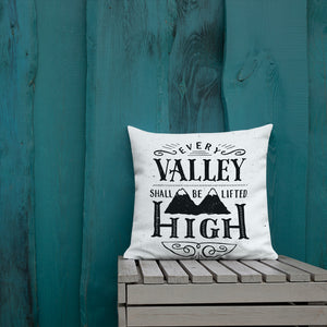 A monochrome square pillow sits on a painted wooden crate against a teal wooden fence. The pillow design is black on a white background, and reads 'Every valley shall be lifted high' in a variety of typographic lettering, with flourishes and an illustration oaif two mountain peaks. 