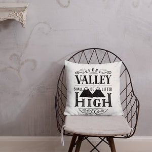 A monochrome square pillow sits on a wire framed chair against a pale grey textured wall. The pillow design is black on a white background, and reads 'Every valley shall be lifted high' in a variety of typographic lettering, with flourishes and an illustration of two mountain peaks. 