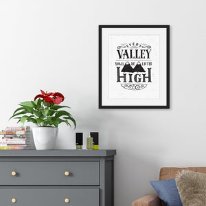 A monochrome black and white print in a black frame hangs on a light wall. The print reads 'Every valley shall be lifted high' in a variety of typographic lettering, with flourishes and an image of two mountain peaks. Beneath the print there’s a grey chest of drawers with a potted plant and books on top, and a brown sofa.