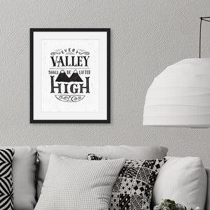 A monochrome black and white print in a black frame hangs on a textured grey living room wall. The print reads 'Every valley shall be lifted high' in a variety of typographic lettering, with flourishes and an image of two mountain peaks. Beneath the print there’s a grey sofa with black and white cushions and a large white lamp.