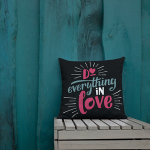 A square black pillow leans against a painted teal fence. The pillow reads "Do everything in love" in bright pink and blue hand-lettering style, with white dashes around the words.