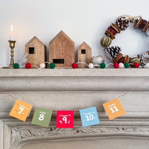 A picture of a fireplace mantle with a candle lit, small houses and a Christmas wreath. Hanging on the mantle on a string are the advent calendar cards showing the numbers of the days in December.