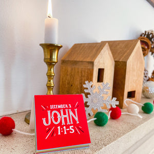 A close up photo of one of the Advent cards showing the Bible verse. In the background on the mantle is a candle and some Christmas decor. 