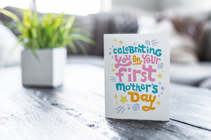 A greeting card featured on a black, wood coffee table. There’s a white planter in the background with a green plant. There’s also a gray sofa in the background with a white pillow. The card features the words “Celebrating you on your first Mother’s Day.”
