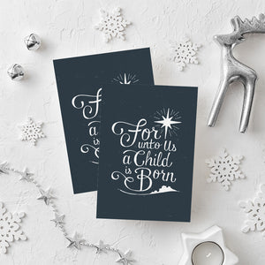 Two Christmas cards laying on a white background with white and silver Christmas decorations on the table. The Christmas card has a navy color as the background with white words reading "for unto us a child is born." Above the words is an illustrated Bethlehem star and below the words is a small illustration of the city of Bethlehem.