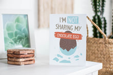 Load image into Gallery viewer, A greeting card featured standing up on a white tabletop with a framed photo of a succulent in the background and a stack of wooden coasters. There’s a woven basket in the background with a cactus inside. The card features an illustrated chocolate Easter egg with the words “I’m not sharing my chocolate egg!”