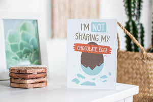 A greeting card featured standing up on a white tabletop with a framed photo of a succulent in the background and a stack of wooden coasters. There’s a woven basket in the background with a cactus inside. The card features an illustrated chocolate Easter egg with the words “I’m not sharing my chocolate egg!”