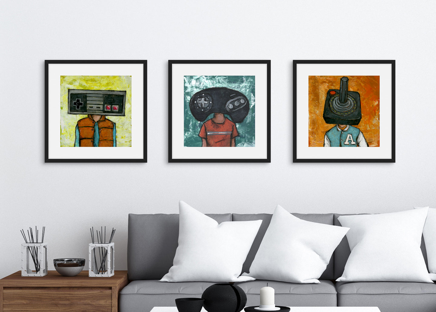 Three black frames on a wall above a grey sofa. The three frames have art prints of illustrated game controllers. The first frame features a vintage Nintendo controller, the second frame a Sega Genesis original controller, and the third frame is an Atari frame. 