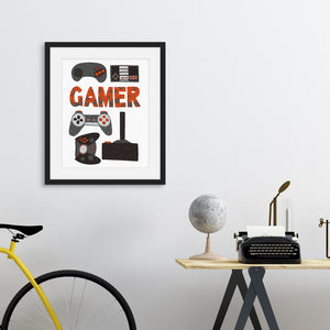 Artwork in a black frame featured in a room with hand drawn lettering and illustrations with different game controllers and the word "gamer." The illustrations and gamer word are in red, grey and black. 