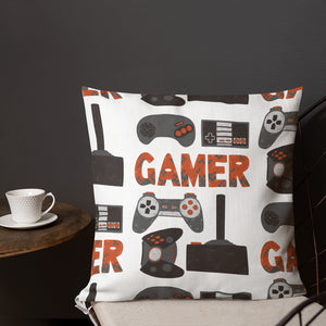 A pillow on a chair with a coffee mug on a table next to it. The white pillow features hand drawn lettering and illustrations featuring different game controllers and the word "gamer." The illustrations and gamer word are in red, grey and black. 
