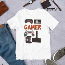 Load image into Gallery viewer, A short sleeved T-shirt laying flat with objects around it. The tee is white and features hand drawn lettering and illustrations featuring different game controllers and the word &quot;gamer.&quot; The illustrations and gamer word are in red, grey and black. 