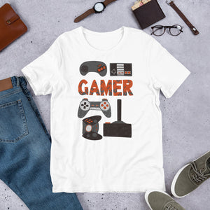 A short sleeved T-shirt laying flat with objects around it. The tee is white and features hand drawn lettering and illustrations featuring different game controllers and the word "gamer." The illustrations and gamer word are in red, grey and black. 