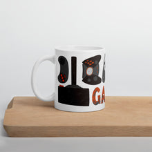 Load image into Gallery viewer, A white mug sitting on a piece of wood. The white mug features hand illustrated images of game controllers and the word gamer in black, red and grey. 