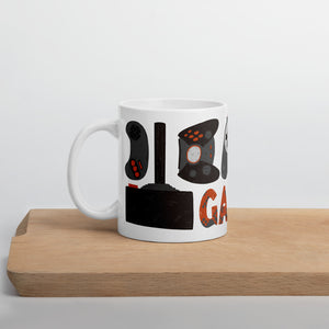 A white mug sitting on a piece of wood. The white mug features hand illustrated images of game controllers and the word gamer in black, red and grey. 