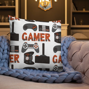 A white pillow on a sofa with a blue knitted blanket with the pillow featuring hand drawn lettering and illustrations featuring different game controllers and the word "gamer." The illustrations and gamer word are in red, grey and black. 