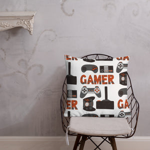 A pillow on a chair against a grey wall. The pillow is white features hand drawn lettering and illustrations featuring different game controllers and the word "gamer." The illustrations and gamer word are in red, grey and black. 