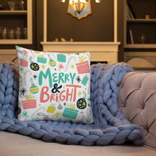Load image into Gallery viewer, A white pillow on a sofa with a blue knitted blanket. The white pillow features a Christmas pattern. The pattern includes the words Merry &amp; Bright in the middle with an illustrated pattern around the words with presents, ornaments, starts and candy canes. The colors of the words and patterns are in pink, light blue, yellow and black.