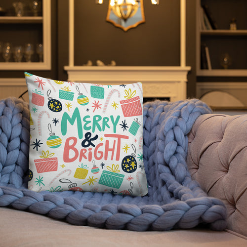 A white pillow on a sofa with a blue knitted blanket. The white pillow features a Christmas pattern. The pattern includes the words Merry & Bright in the middle with an illustrated pattern around the words with presents, ornaments, starts and candy canes. The colors of the words and patterns are in pink, light blue, yellow and black.