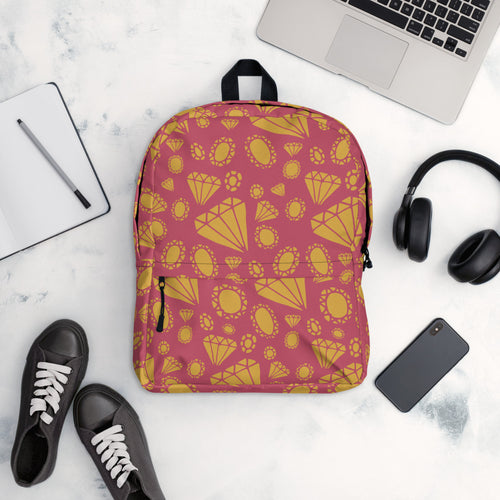 A backpack is placed on a table with a laptop, notebook, shoes, headphones and a mobile phone. The backpack features an illustrated design of gems and diamonds in gold. The gem pattern is on top of a muted hot pink color. 
