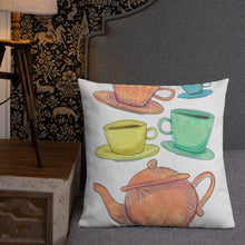 Load image into Gallery viewer, A pillow leaning on a grey headboard with a plant in the background. A pillow on a chair against a grey wall. The white pillow features the artwork on a white background with four teacups on saucers and one large teapot. The teacups are in muted colors of orange, blue, yellow and green and the teapot is a muted orange. On the teacups, saucers and teapot there is a light flower detail pattern.