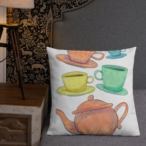 A pillow leaning on a grey headboard with a plant in the background. A pillow on a chair against a grey wall. The white pillow features the artwork on a white background with four teacups on saucers and one large teapot. The teacups are in muted colors of orange, blue, yellow and green and the teapot is a muted orange. On the teacups, saucers and teapot there is a light flower detail pattern.
