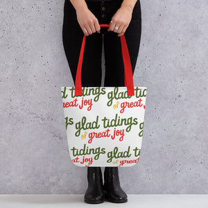 Someone holding a tote bag with red handles and a white fabric bag. The tote bag features the words "glad tidings of great joy" in green, yellow and red. 