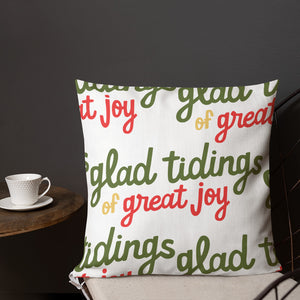 A pillow on a chair with a coffee mug on a table next to it. The lettering says "glad tidings of great joy" in red, green and yellow. The words create a pattern on the pillow. 