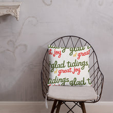 Load image into Gallery viewer, The pillow is leaning on a metal chair with a cushion. The white pillow has a repeat pattern with the phrase &quot;glad tidings of great joy&quot; in the colors green, red and yellow. 