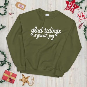 An olive green sweatshirt laying on a table with Christmas objects around it. The sweatshirt has the words "glad tidings of great joy" in white. 